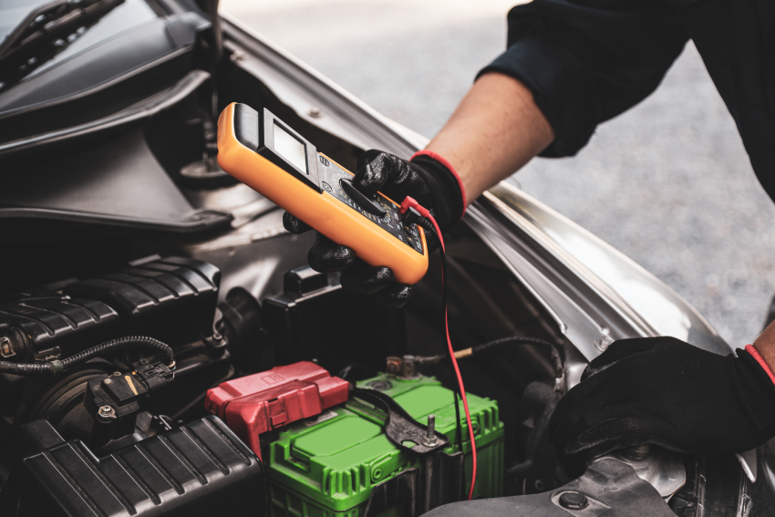 Person using an orange multimeter to check a car battery. The hood is open and the person's hands are wearing black gloves.