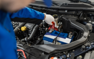 Mechanic in blue uniform connects a red jumper cable to a car battery under the hood.