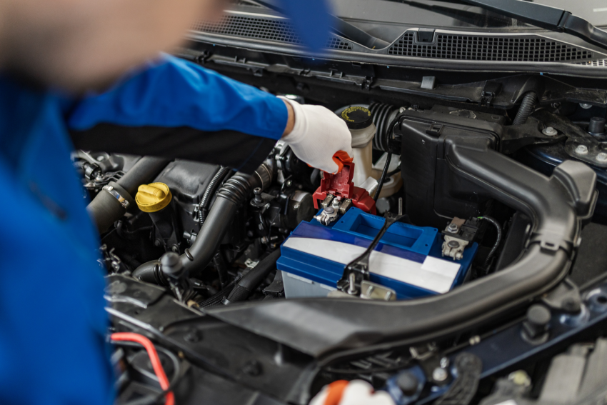 Mechanic in blue uniform connects a red jumper cable to a car battery under the hood.