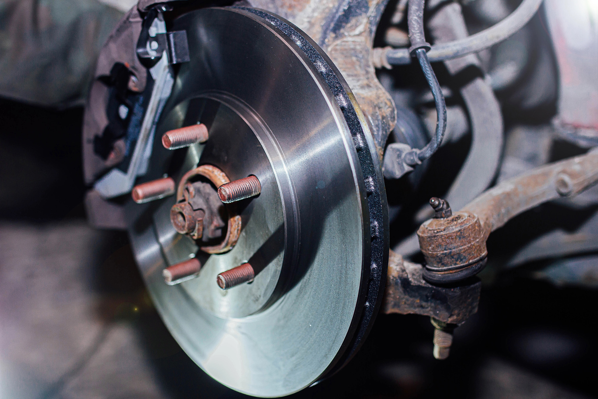 Close-up of a car's brake disc and wheel hub assembly, illustrating four rusty lug bolts, the shiny metal rotor, and surrounding suspension components—a reminder that timely brake service can keep these parts in optimal condition.