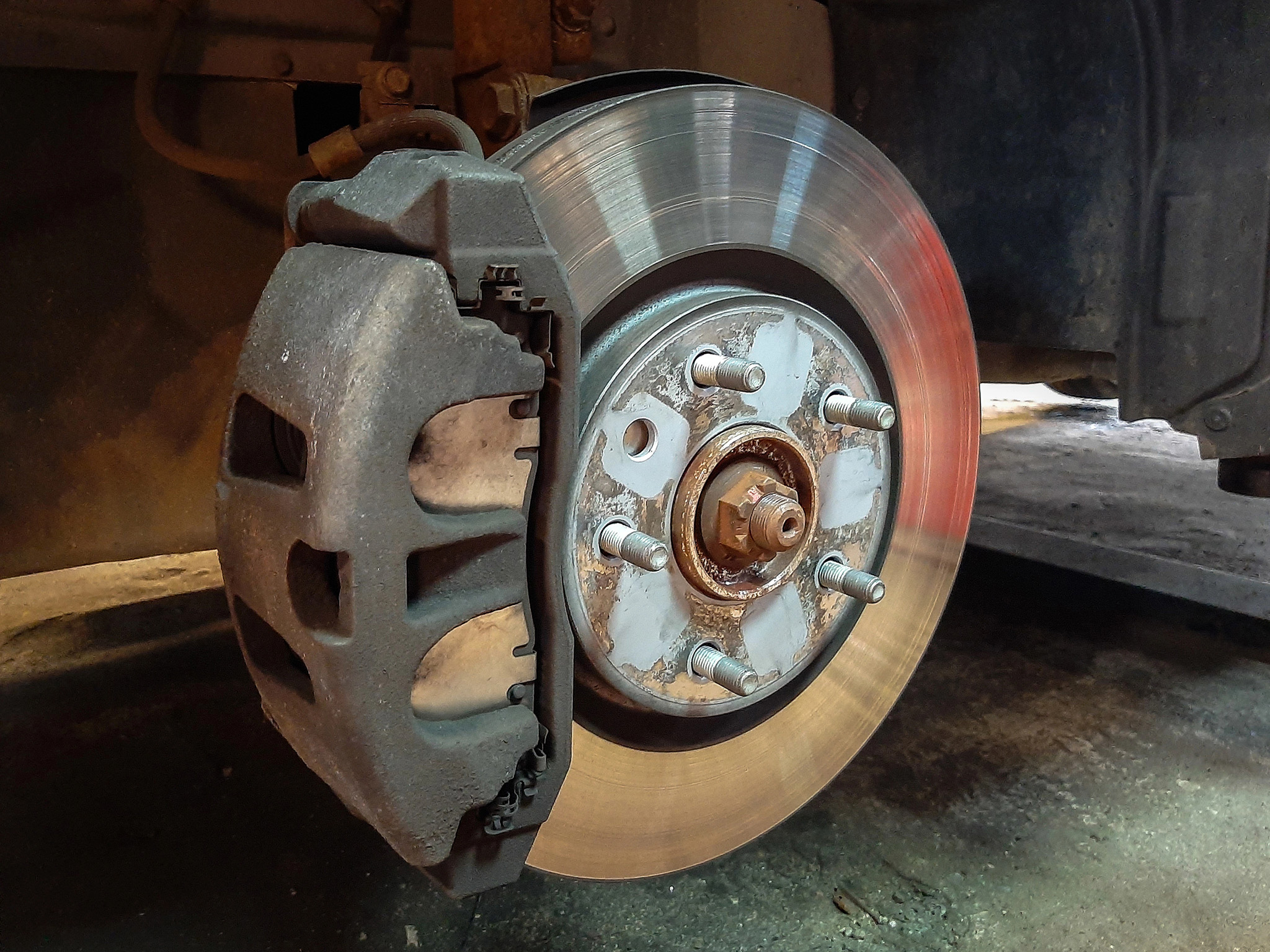 Close-up of a car's exposed disc brake and caliper, with six lug nuts visible, set against a vehicle's wheel hub in a garage environment, highlighting the importance of regular brake service.