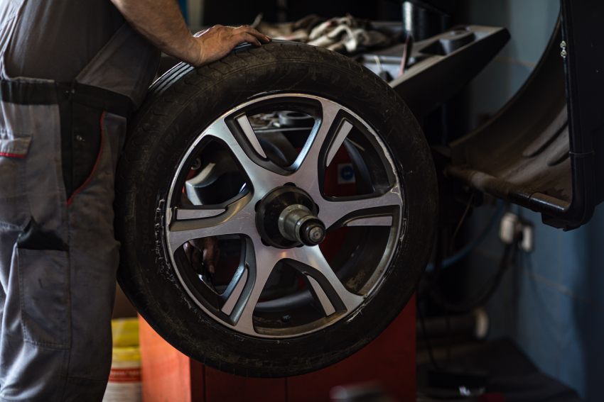 Mechanic stands next to a tire mounted on a balancing machine, with tools and equipment visible in the background.