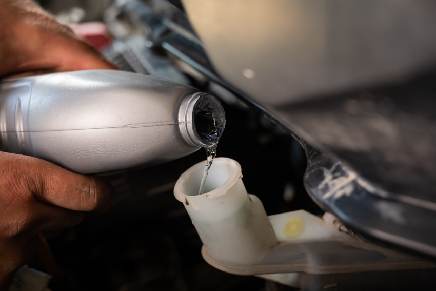 Person pouring liquid from a gray bottle into an engine reservoir under a car hood.