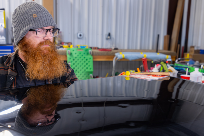 A person with a long red beard and glasses wearing a gray beanie and plaid shirt looks at a polished car surface in a garage, with tools and equipment in the background.