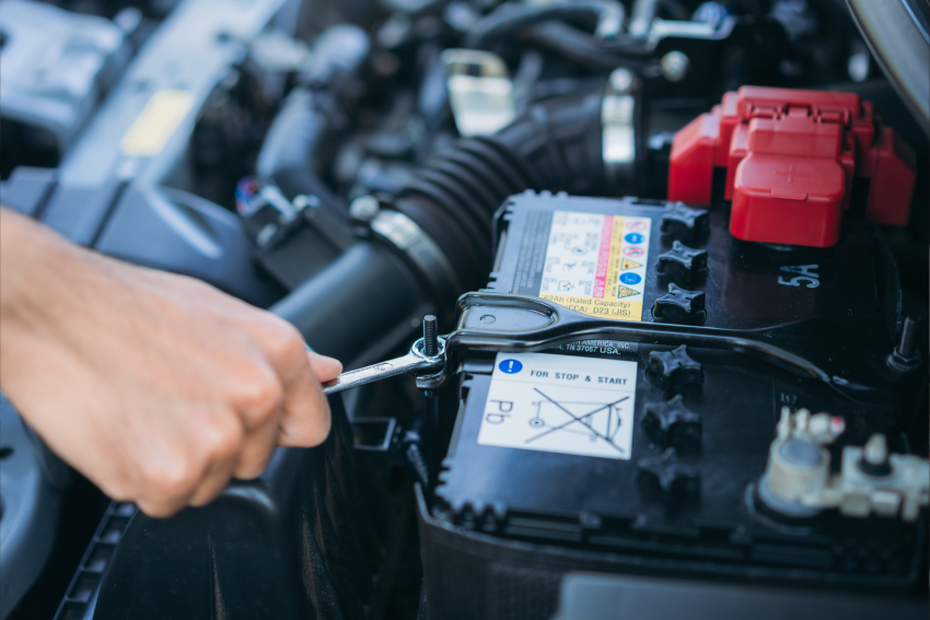 A hand with a wrench tightens a bolt on a car battery under the hood.