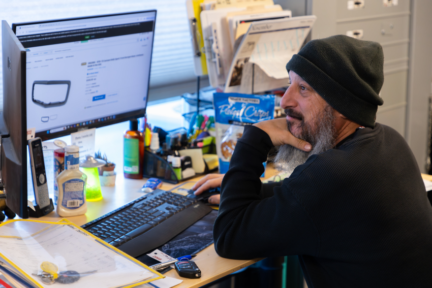 Matt wearing a beanie is seated at a cluttered desk, looking at a computer screen displaying a product. Office supplies and papers are scattered around.