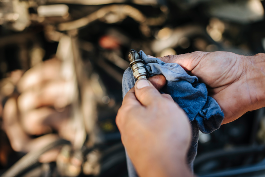 Hands cleaning a spark plug with a blue cloth in front of a car engine.