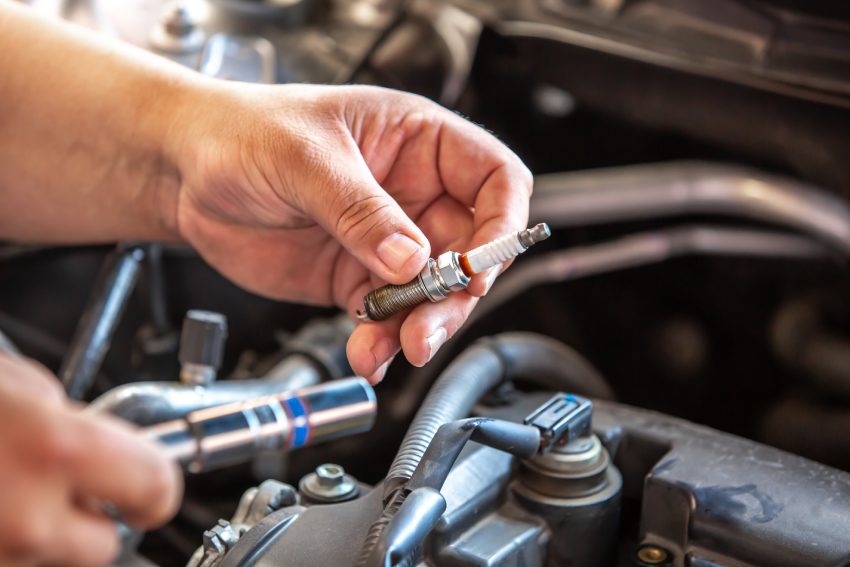 Hands holding a spark plug over an open car engine, surrounded by mechanical components and tools.