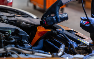 Person wearing black gloves pouring oil into a car engine using a funnel in a garage.
