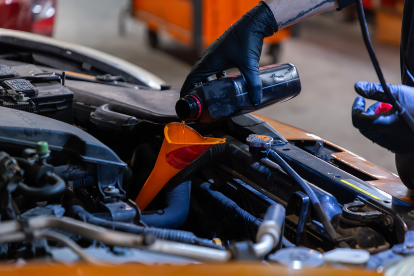 Person wearing black gloves pouring oil into a car engine using a funnel in a garage.