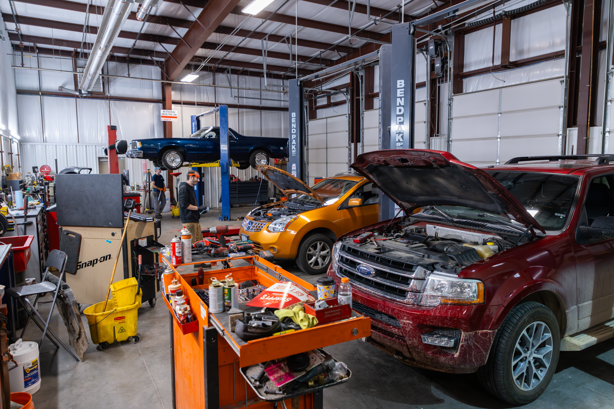 Auto repair shop with three cars being serviced. The red SUV has its hood open, an orange car is on the ground, and a blue car is elevated on a lift. Tools are scattered around.