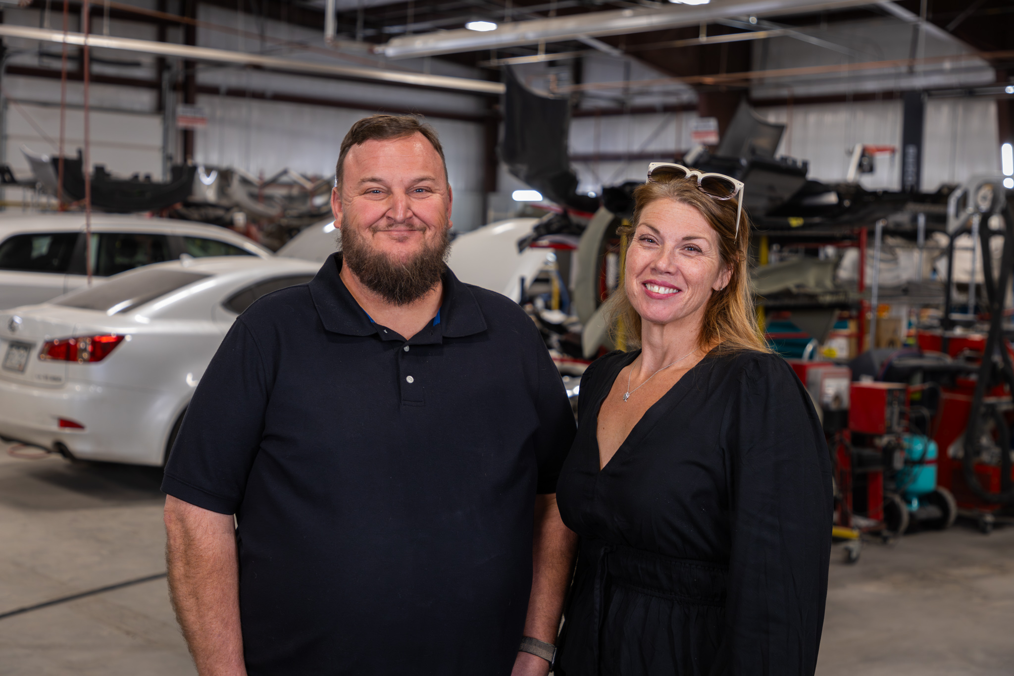 Two people standing in an auto repair shop, smiling at the camera.