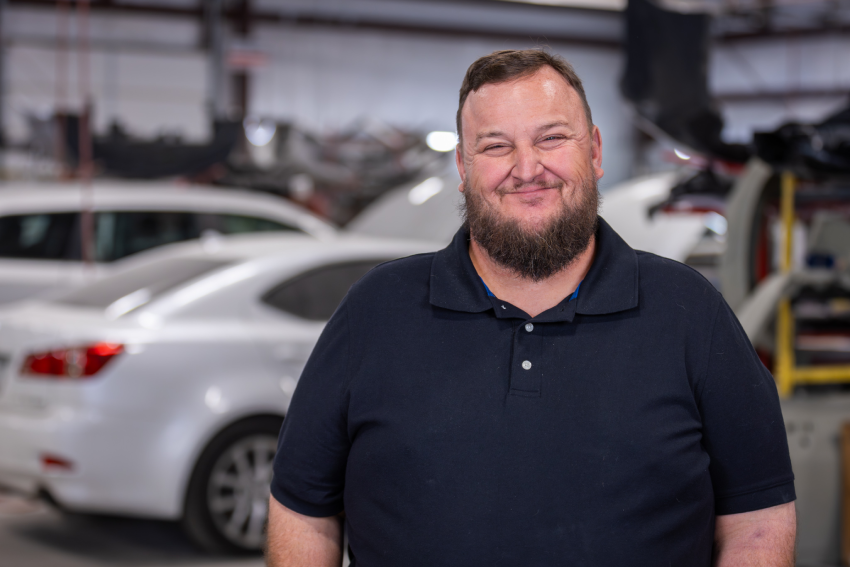 Bill Ellison in a black polo shirt stands smiling in an auto repair shop, with white cars and various tools in the background.