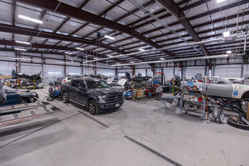 Interior of an auto repair shop with various vehicles and equipment. A pickup truck is parked in the center. The space is well-lit, with a high ceiling and exposed metal beams.