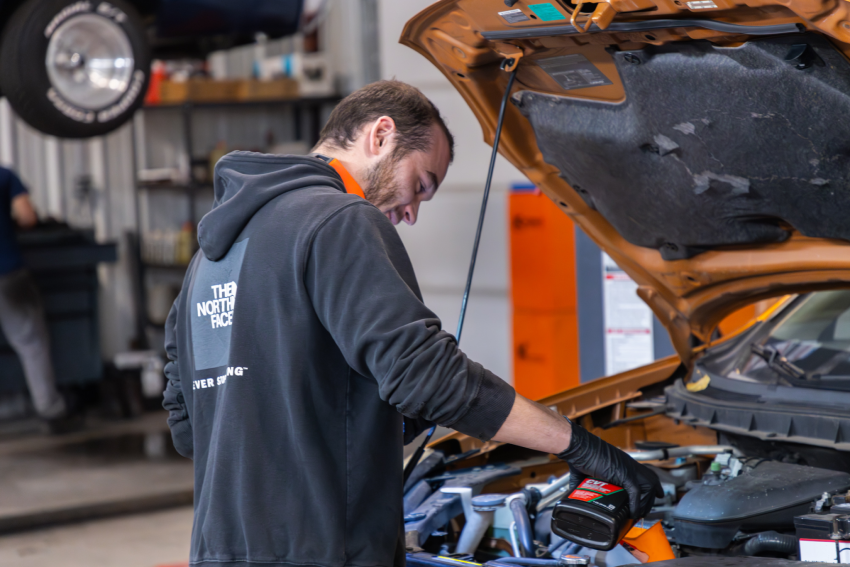 A man in a black hoodie and gloves changes the oil of a car with an open hood in a garage setting.