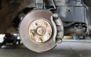 Close-up of a car disc brake with visible rust and wear, mounted on the wheel hub at an auto service shop. The surrounding area reveals part of the vehicle's suspension system.