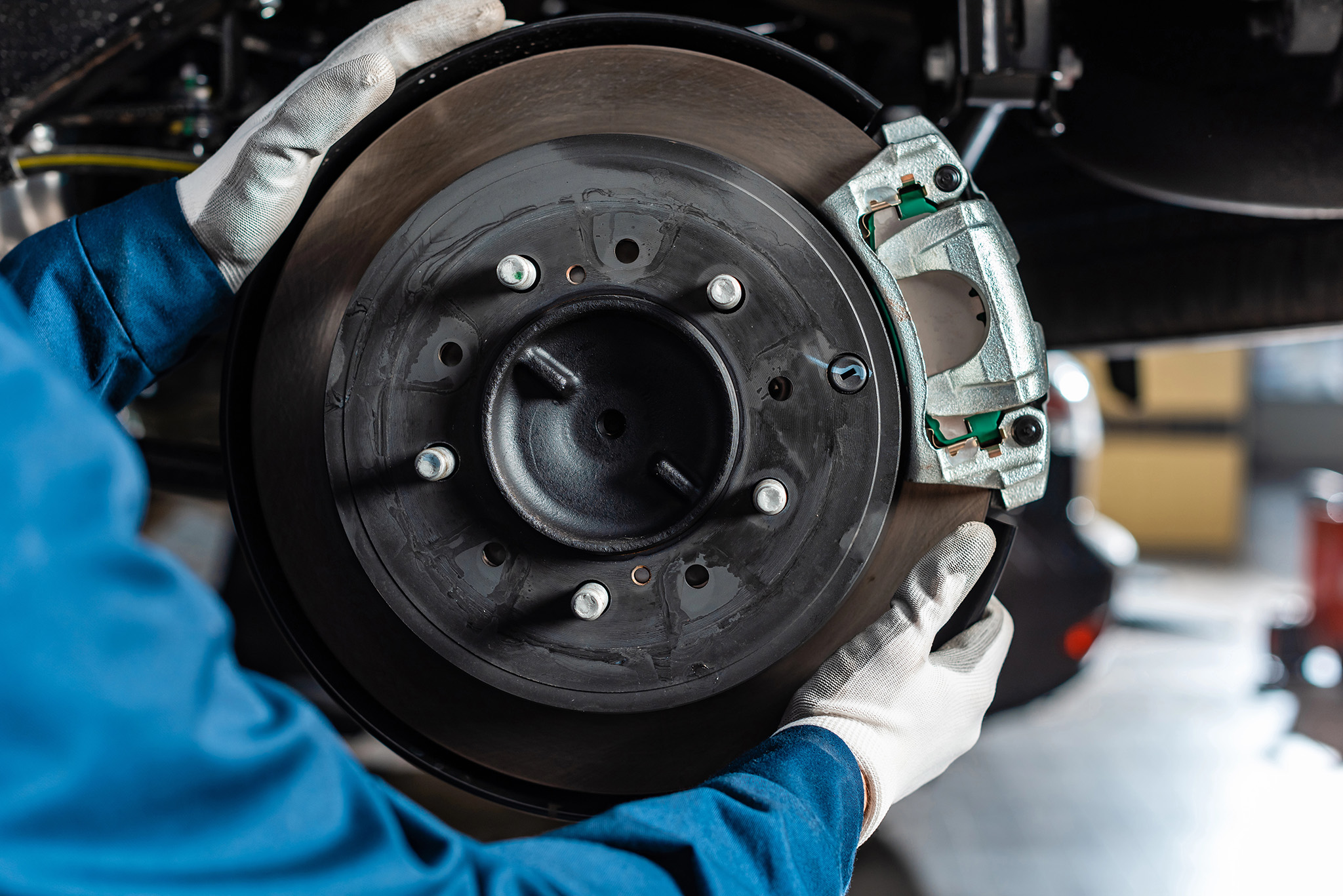 In an auto service shop, a mechanic in blue overalls and white gloves diligently works on a car's brake disc and caliper, surrounded by tools in the bustling garage setting.