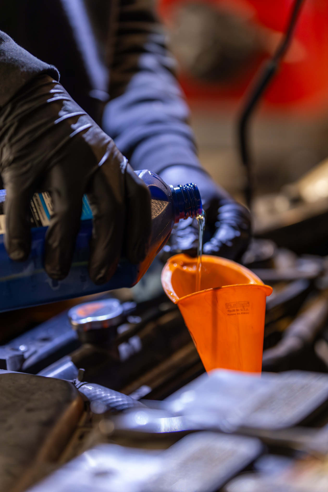 In an auto service shop, a person wearing gloves carefully pours blue liquid into a car engine using an orange funnel.