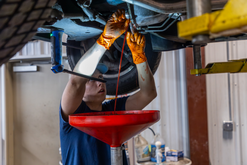 Mechanic wearing orange gloves under a car, using a tool as red fluid drains into a red funnel.