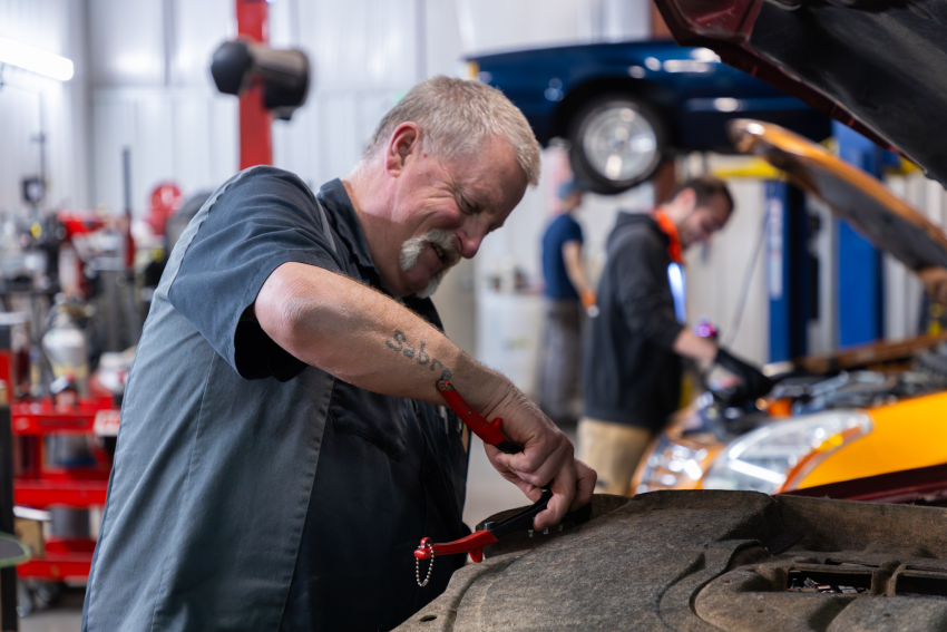 Mechanic working on car engine with a tool in a garage. Another person and vehicles in the background.