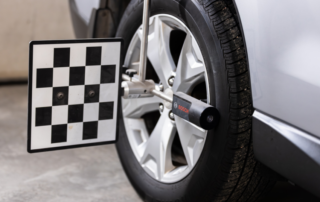 Car wheel alignment tool attached to a vehicle's front tire, with a checked calibration target plate and alignment equipment visible.