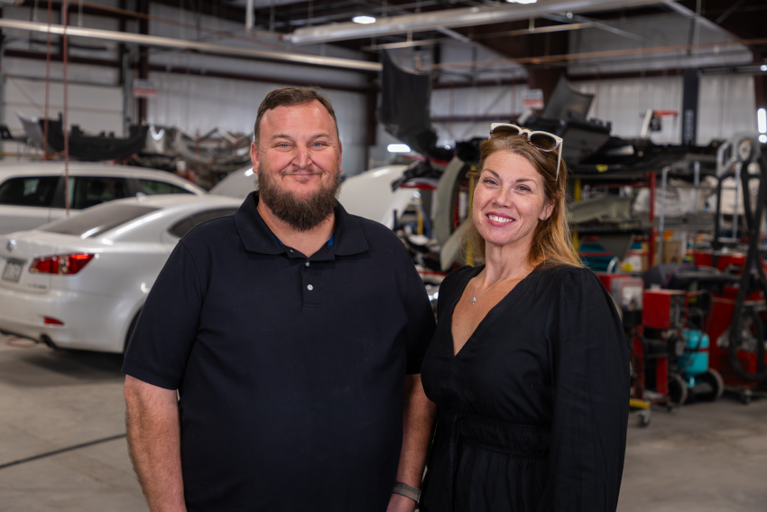 Bill & Stephanie Ellison, Owners of Tri-Lakes Service Center, smiling in an auto body shop, with cars and repair equipment visible in the background.