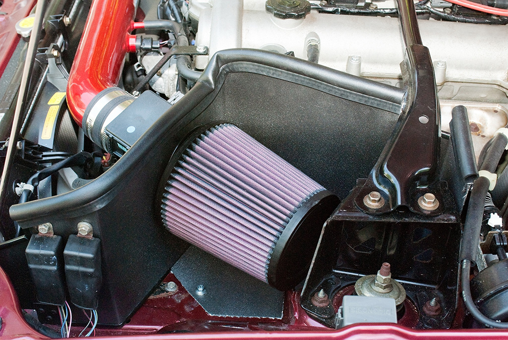 Close-up of a car engine with a modified air intake system, ideal for those considering engine swaps. The cylindrical, pleated air filter is securely attached to a striking red intake pipe.