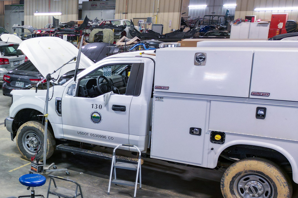 In a tire shop, a white utility truck with its hood open is being worked on. Various tools and other vehicles are visible in the background, highlighting the hive of activity.