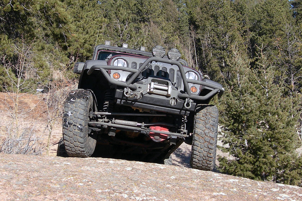 A rugged off-road vehicle, fresh from the tire shop, climbs a rocky incline in a forested area, displaying its suspension and front bumper.