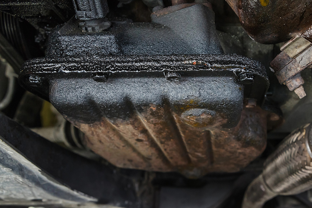 Close-up of a rusted and oil-stained car engine oil pan visible from the underside of a vehicle, surrounded by mechanical components in need of engine repair.