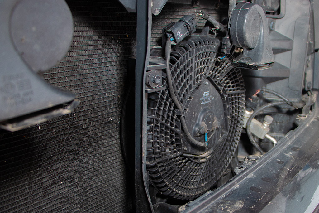 Close-up of a car radiator fan and shroud inside an engine compartment, ready for a fluid flush. The fan is encased in a circular, grid-patterned cover.