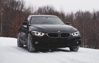 A black BMW sedan with headlights on is parked on a snowy road, surrounded by leafless trees, during active snowfall.
