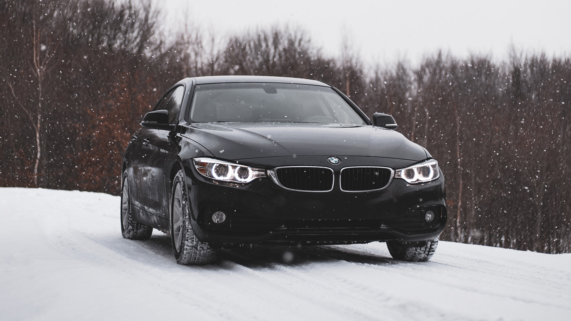 A black BMW sedan with headlights on is parked on a snowy road, surrounded by leafless trees, during active snowfall.